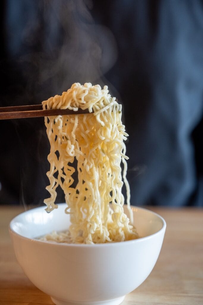 A close-up of the Ve Wong Kung-Fu Chicken Flavor Instant Noodle being lifted from a white bowl with wooden chopsticks, showing thin wavy pale wheat noodles with a slight yellow tint and steam rising above them.