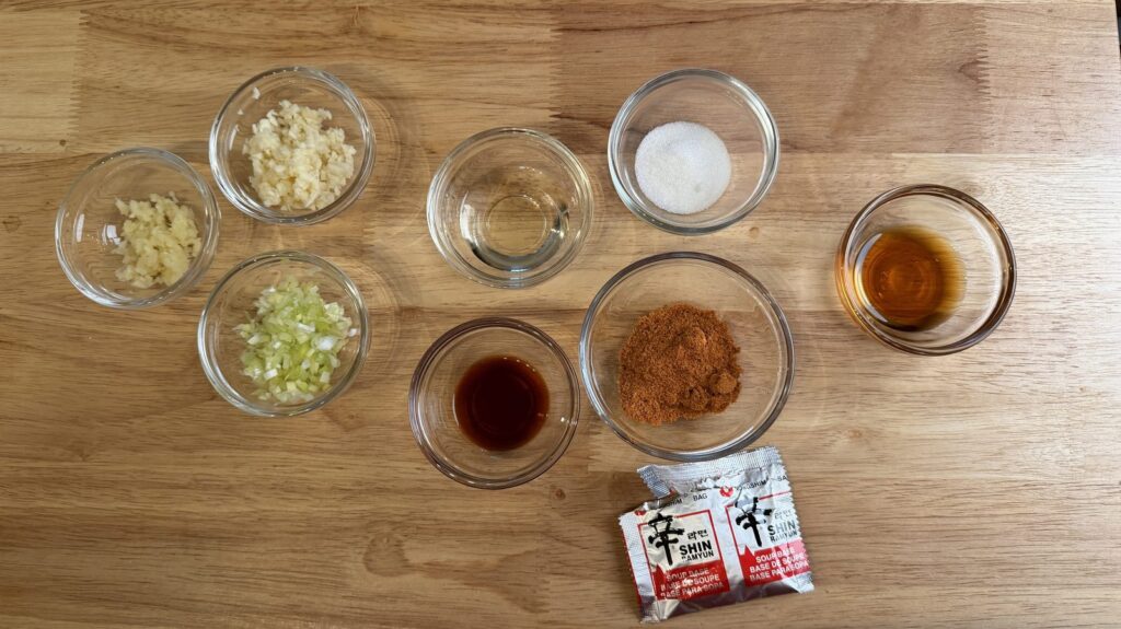 Mise en place for Shin Ramyun rice bowl: glass bowls of garlic, ginger, scallion whites, soy sauce, mirin, sugar, sesame oil, and one Shin Ramyun seasoning packet