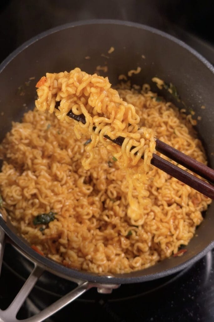 Chopsticks lifting seasoned Shin Ramyun noodles from a pot during the stir-fry step