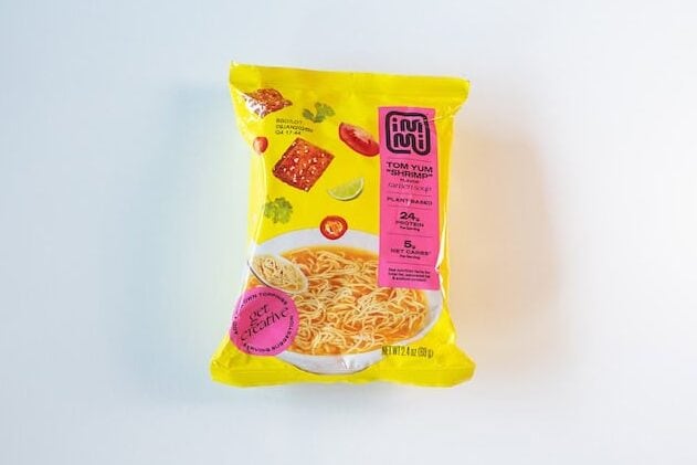 The yellow package of Immi Tom Yum Shrimp Ramen on a white surface, showing illustrated toppings (a halved tofu cube with sesame seeds, sliced red chili, lime wedge, cilantro, tomato slice) above an illustrated bowl of noodles, with a hot pink TOM YUM SHRIMP stripe down the right side and 24g PROTEIN / 5g NET CARBS in a pink banner.