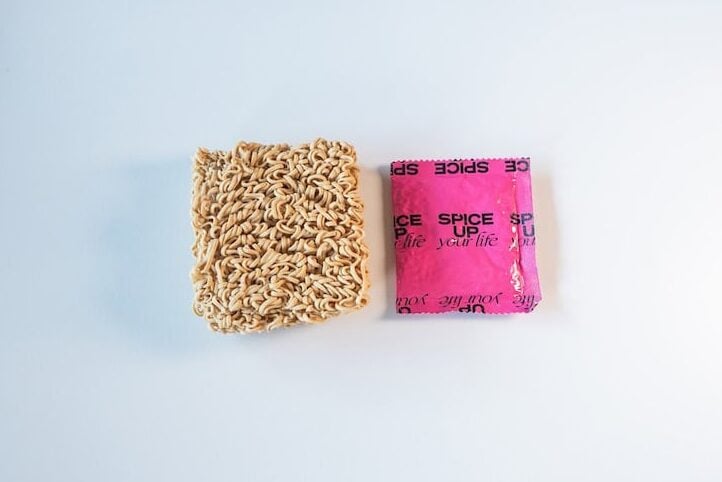 The contents of the Immi Tom Yum Shrimp Ramen pack laid out on a white surface, showing a small dense rectangular block of thin wavy plant-based noodles next to a hot pink Immi soup base sachet that reads SPICE UP your life.