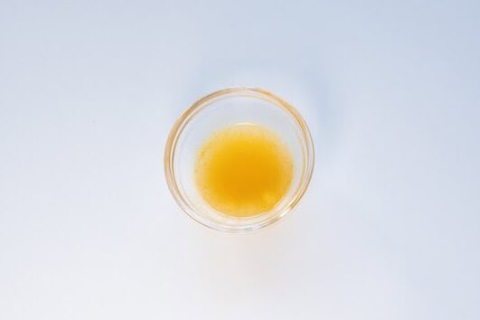 A small clear glass ramekin of the Immi Tom Yum Shrimp Ramen broth on a white surface, showing a thin, golden-orange liquid.