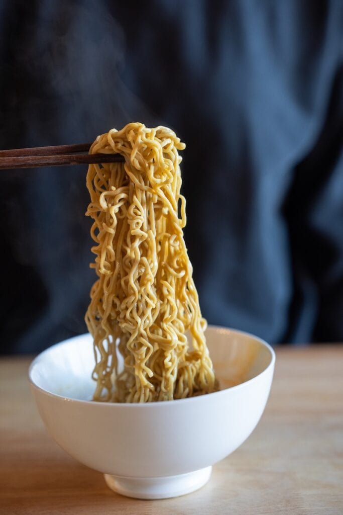 A close-up of the Immi Tom Yum Shrimp Ramen being lifted from a white bowl with wooden chopsticks, showing thin pale wavy plant-based noodles coated in a glossy golden tom yum broth with steam rising above them.