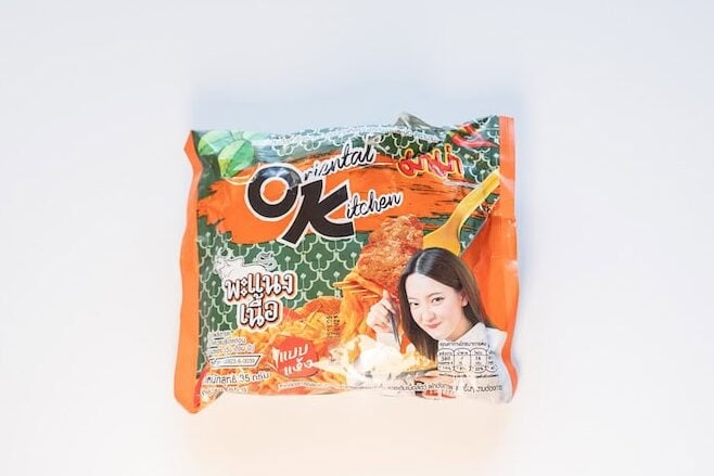 The orange and green package of MAMA Oriental Kitchen Instant Noodles Beef Panang Curry on a white surface, showing the OK Oriental Kitchen logo at the top left, the MAMA brand mark at the top right, and a model holding chopsticks of finished orange-red noodles next to the Thai product name in red script.