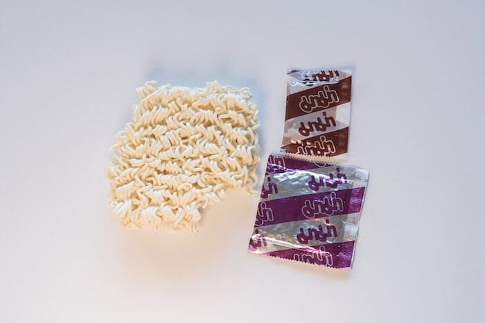 The contents of the MAMA Oriental Kitchen Instant Noodles Beef Panang Curry pack laid out on a white surface, showing a square block of thin wavy wheat noodles on the left and two foil sachets stacked on the right, a brown sauce packet on top and a larger purple seasoning powder packet below.