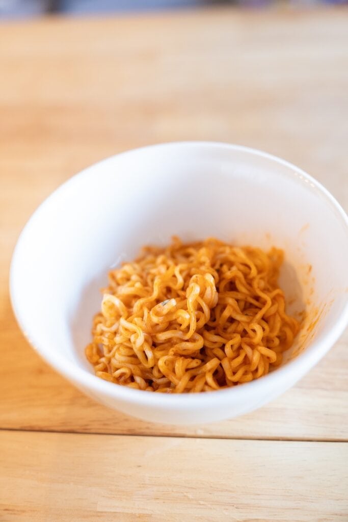 A white bowl of the finished MAMA Oriental Kitchen Instant Noodles Beef Panang Curry on a wooden table, showing thin wavy wheat noodles coated in an orange-red Panang curry sauce.
