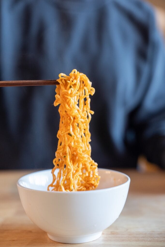 A close-up of the MAMA Oriental Kitchen Instant Noodles Beef Panang Curry being lifted from a white bowl with wooden chopsticks, showing thin wavy wheat noodles glossy with orange-red Panang curry sauce.