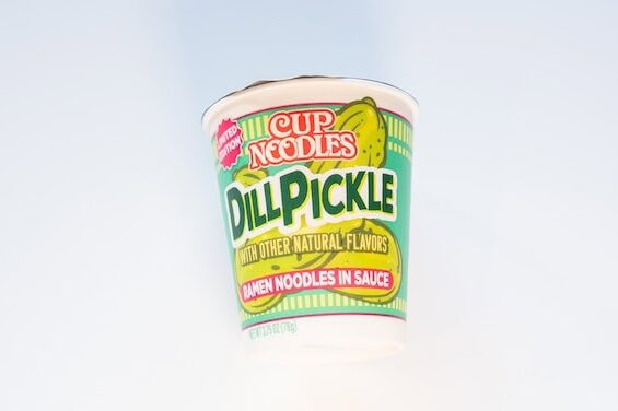 The bright green and pink cup of Nissin Cup Noodles Dill Pickle on a white surface, showing the red LIMITED EDITION badge in the upper left, the red CUP NOODLES bubble logo at the top, DILL PICKLE in large dark green text with illustrated pickle slices behind it, WITH OTHER NATURAL FLAVORS underneath, and a pink banner reading RAMEN NOODLES IN SAUCE