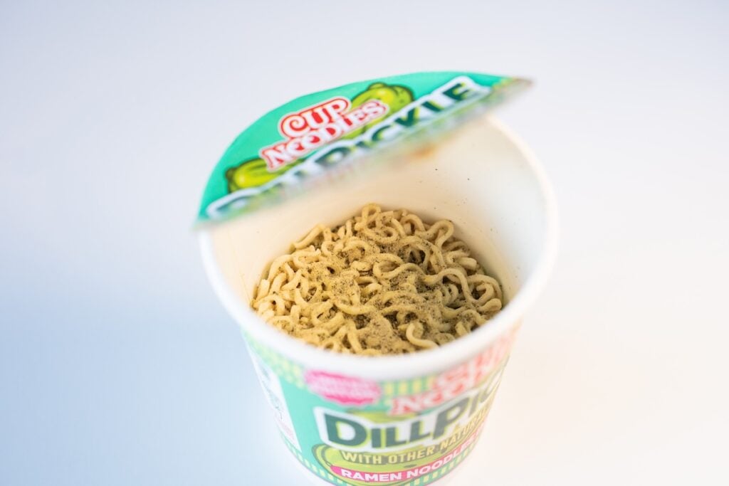 The contents of the Nissin Cup Noodles Dill Pickle cup with the paper lid partially peeled back, showing the fried wavy noodle brick dusted with a fine green-grey dill pickle seasoning powder that also coats the inside walls of the cup.
