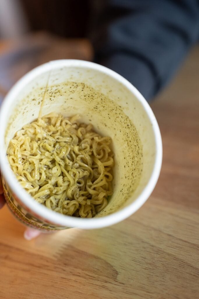 A top-down view of the finished Nissin Cup Noodles Dill Pickle in its paper cup held at an angle, showing thin wavy fried wheat noodles coated in a glossy green-grey dill pickle seasoning, with the inside wall of the cup dusted with more of the powder.