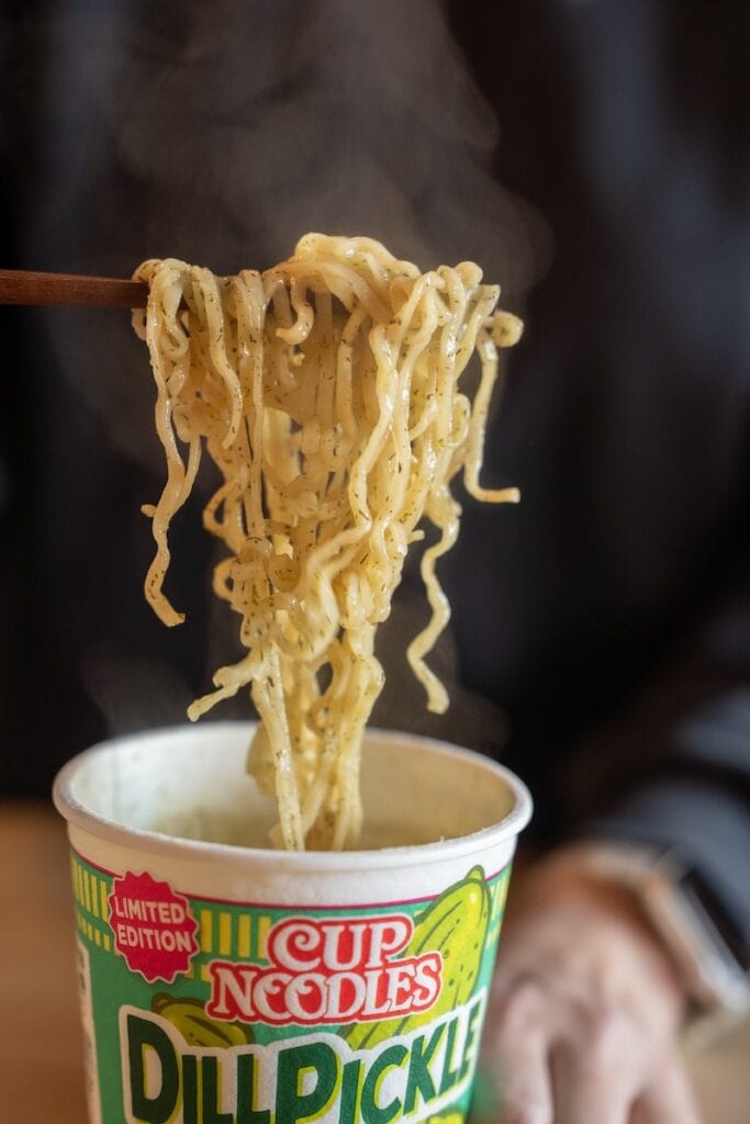 A close-up of the Nissin Cup Noodles Dill Pickle being lifted from the paper cup with wooden chopsticks, showing thin wavy fried wheat noodles coated in green dill pickle seasoning with steam rising above them.