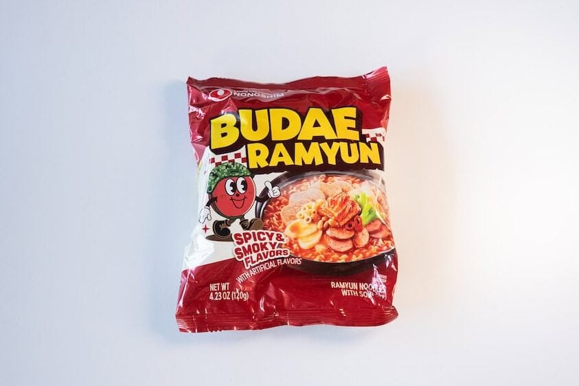 The deep red package of Nongshim Budae Ramyun Noodles with Soup Mix on a white surface, showing the white NONGSHIM logo at the top, BUDAE RAMYUN in large yellow lettering, a small red Budae mascot character, SPICY & SMOKY FLAVORS in white inside a red banner, and a photo of finished red Korean army stew noodles topped with sliced sausage, kimchi, green onion, and white pieces of pressed meat.