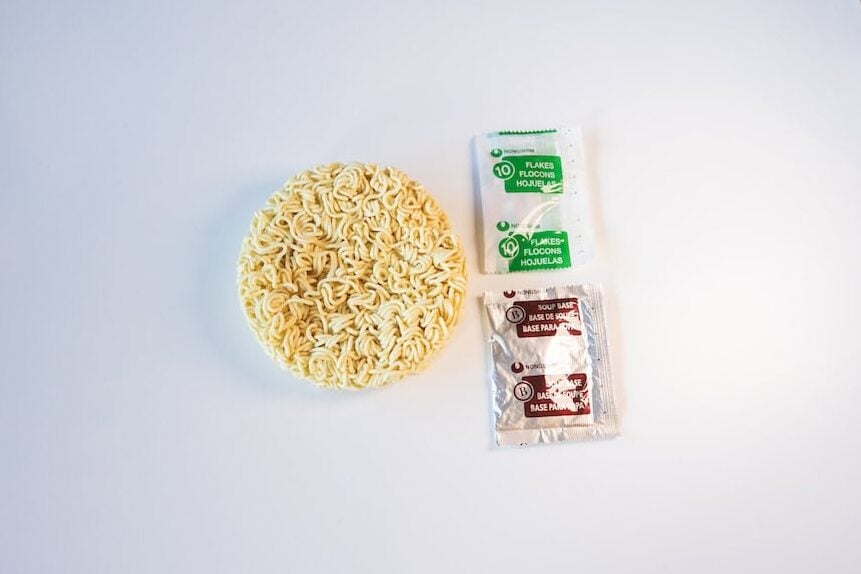 The contents of the Nongshim Budae Ramyun Noodles with Soup Mix pack laid out on a white surface, showing a round disc of fried wavy wheat noodles on the left, a small green Nongshim Flakes packet at the top right, and a larger silver foil Nongshim Soup Base sachet below it.