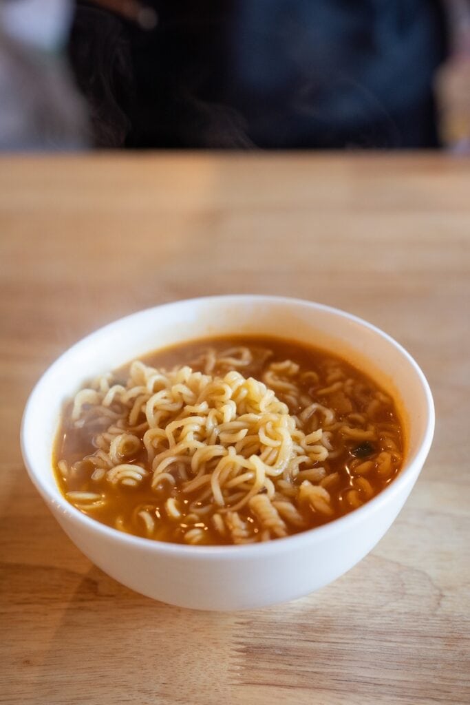 A white bowl of the finished Nongshim Budae Ramyun Noodles with Soup Mix on a wooden table, showing wavy wheat noodles in a deep orange-red spicy broth with visible kimchi flakes and small pieces of soy-protein meat