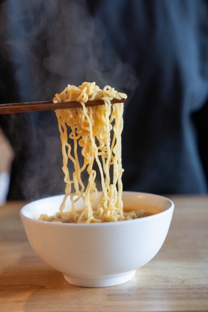 A close-up of the Nongshim Budae Ramyun Noodles with Soup Mix being lifted from a white bowl with wooden chopsticks, showing wavy wheat noodles glossy with red spicy broth and steam rising above them