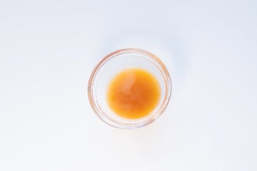 A small clear glass ramekin of the Nongshim Budae Ramyun Noodles with Soup Mix broth on a white surface, showing a deep orange-red liquid with a slight oil sheen.