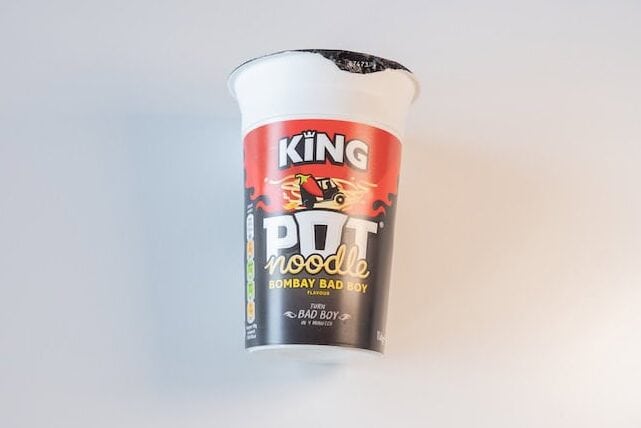 The tall white cup of Pot Noodle King Bombay Bad Boy on a white surface, showing the red KING crown logo at the top, a chili pepper riding a motorcycle illustration, bold white POT noodle lettering, BOMBAY BAD BOY FLAVOUR in white inside a red banner, and a TURN BAD BOY IN 4 MINUTES callout.
