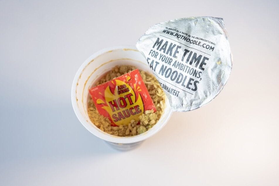 The contents of the Pot Noodle King Bombay Bad Boy cup with the silver foil lid partially peeled back, showing the dry wheat noodle brick dusted with yellow-brown curry seasoning, dehydrated green peas and corn kernels, and a red and yellow HOT SAUCE sachet sitting on top ready to add at the end.