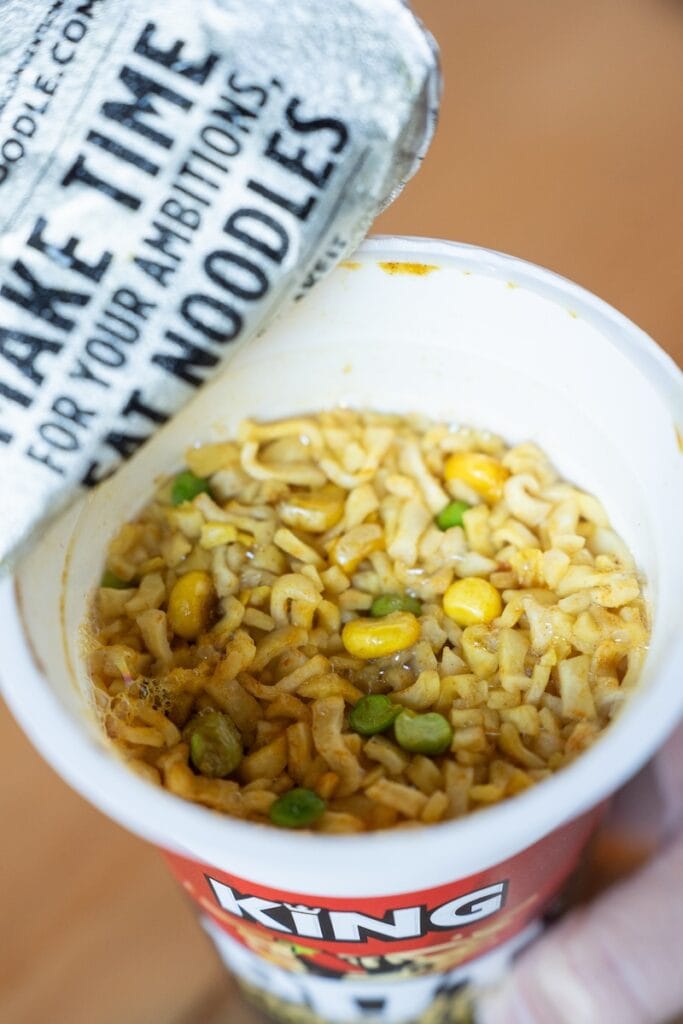 A top-down view of the finished Pot Noodle King Bombay Bad Boy in its tall paper cup, showing thick egg-noodle-style wheat noodles coated in a thick yellow-orange curry sauce with dehydrated corn kernels and green peas scattered throughout.