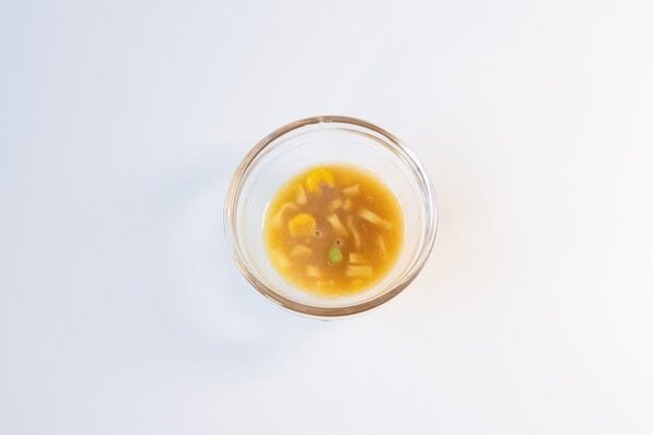 A small clear glass ramekin of the Pot Noodle King Bombay Bad Boy broth on a white surface, showing a thick yellow-orange curry liquid with visible corn kernels and a green pea floating in it.