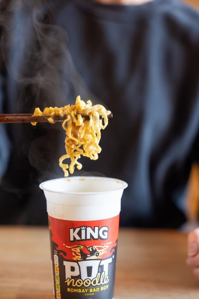 A close-up of the Pot Noodle King Bombay Bad Boy being lifted from the tall paper cup with wooden chopsticks, showing thick egg-noodle-style wheat noodles coated in yellow-orange curry sauce with steam rising above them.