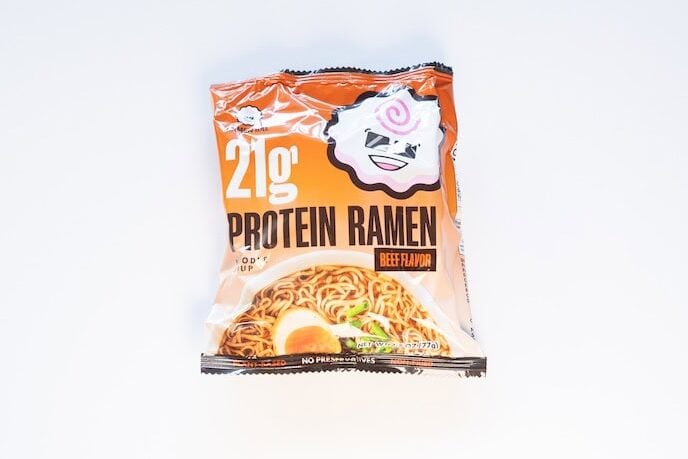 The orange package of Ramen Bae Beef Protein Ramen Soup on a white surface, showing a small white Ramen Bae mascot face at the top, 21g PROTEIN RAMEN in bold black text with a tiny orange BEEF FLAVOR tag, NOODLE SOUP underneath, a photo of finished beef ramen with a halved soft-boiled egg and sliced green onion, and a NO PRESERVATIVES callout at the bottom.