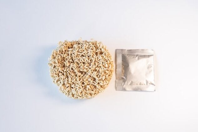 The contents of the Ramen Bae Beef Protein Ramen Soup pack laid out on a white surface, showing a round disc of pale high-protein wavy wheat noodles on the left and a single silver foil seasoning packet on the right.