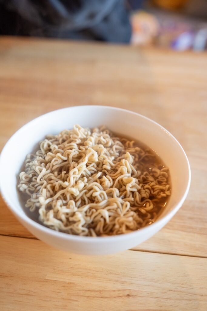 A white bowl of the finished Ramen Bae Beef Protein Ramen Soup on a wooden table, showing wavy pale wheat noodles in a clear amber-brown beef broth.