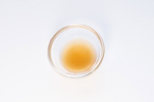 A small clear glass ramekin of the Ramen Bae Beef Protein Ramen Soup broth on a white surface, showing a pale amber-orange liquid with a slight oil sheen.