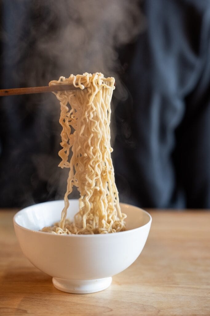 A close-up of the Ramen Bae Beef Protein Ramen Soup being lifted from a white bowl with wooden chopsticks, showing wavy sturdy high-protein wheat noodles with steam rising above them.
