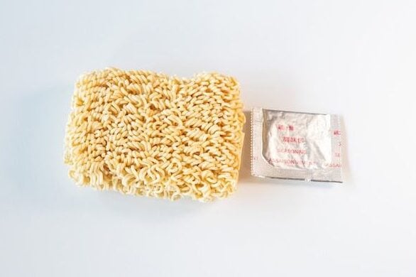 The contents of the Ve Wong Kung-Fu Chicken Flavor pack laid out on a white surface, showing a small rectangular brick of thin wavy pale wheat noodles on the left and a small silver foil Ve Wong Seasoning packet on the right.