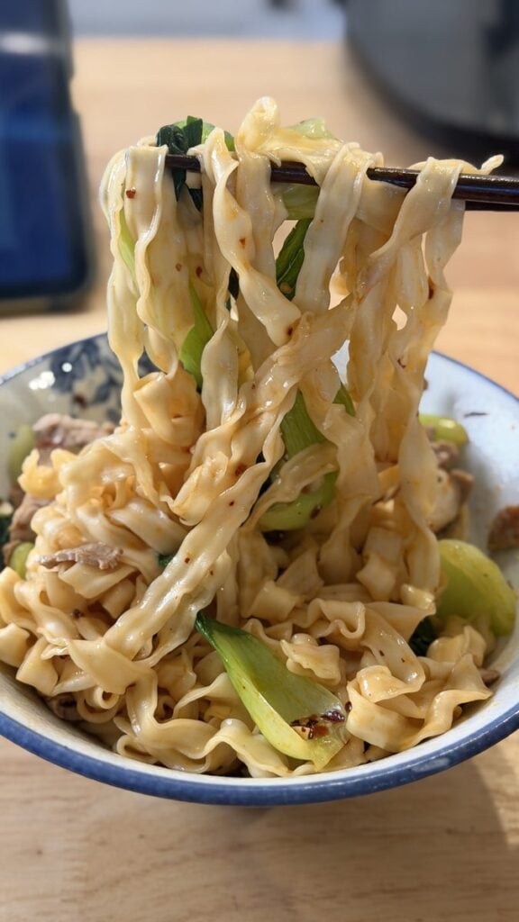 Chopsticks lifting easy chili oil noodles with bok choy and pork loin from a bowl