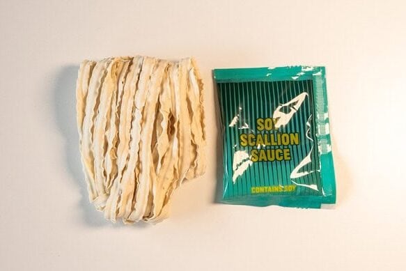 The contents of the Fly By Jing pack on a white surface: a bundle of flat, wavy sun-dried wheat noodles next to a single teal Soy Scallion Sauce packet.
