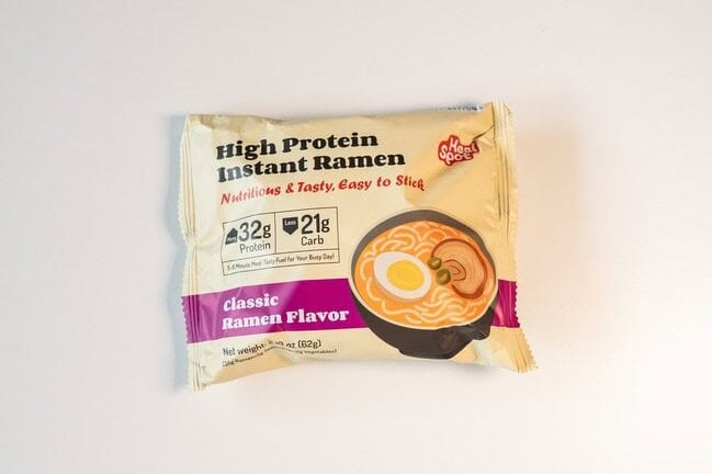 The cream and yellow Heal Spot High Protein Instant Ramen package on a white surface, showing the product name in black, a red Nutritious and Tasty tagline, 32g Protein and 21g Carb badges, and a purple Classic Ramen Flavor banner.