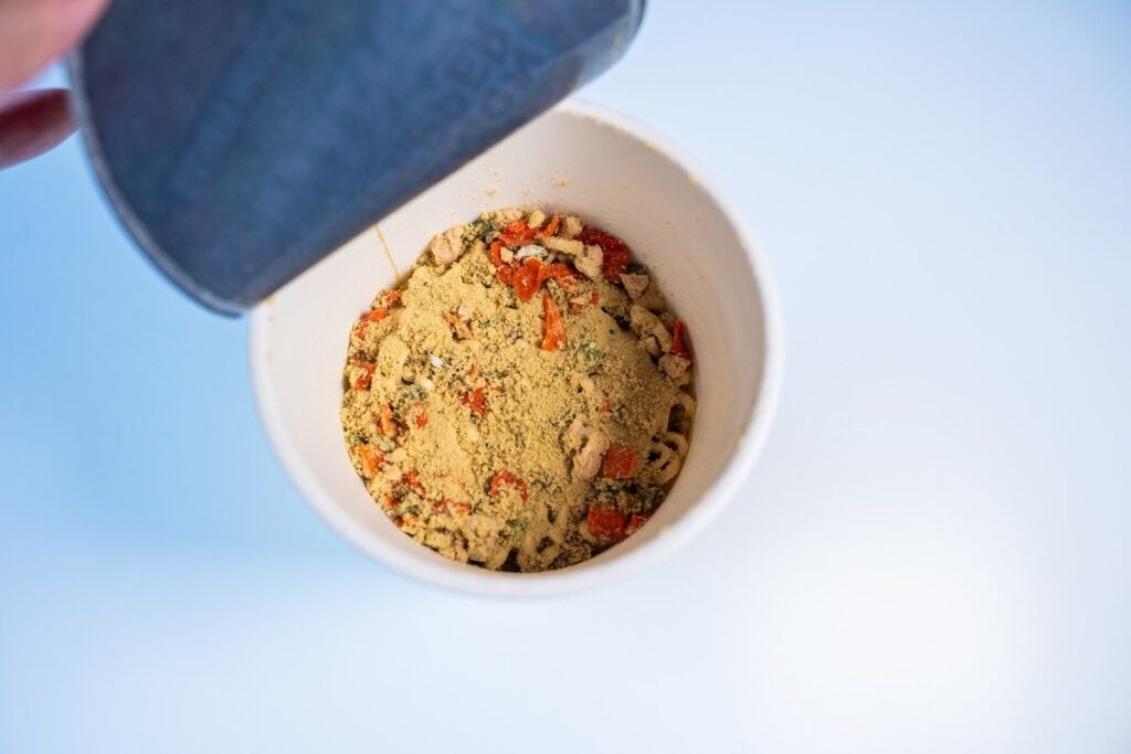 The dry contents of the Nissin Cup Noodles Lemon Pepper Wings Ramen Noodles tipped forward onto the noodle block, showing pale yellow seasoning powder, orange dried carrot pieces, small chunks of textured soy protein, and scattered green herbs with visible cracked black pepper.