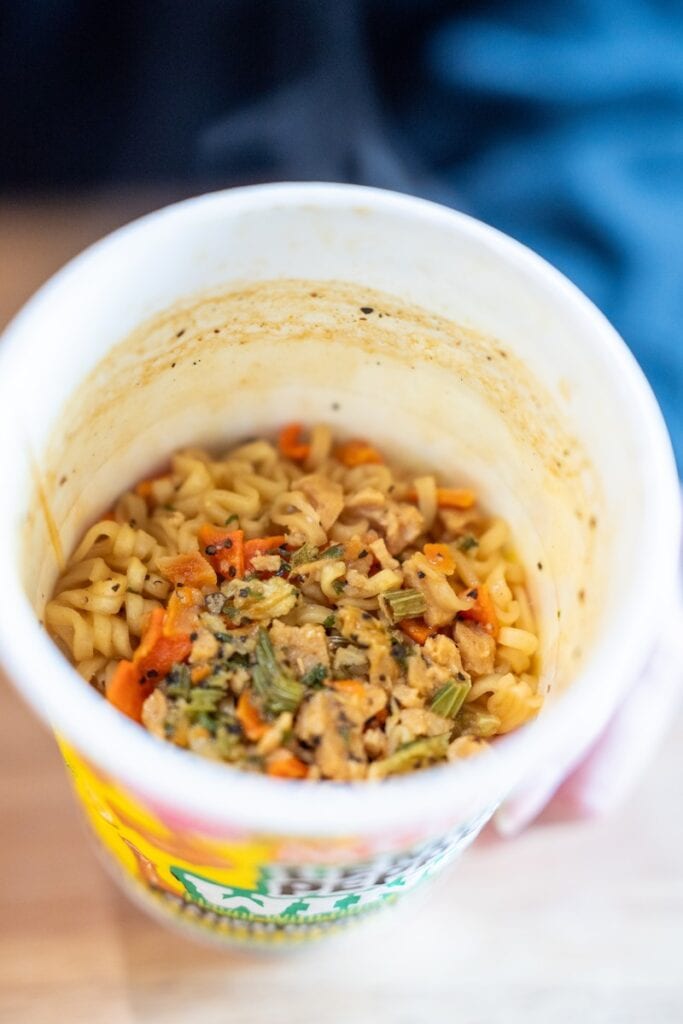 Overhead close-up of the cooked Nissin Cup Noodles Lemon Pepper Wings Ramen Noodles inside the cup, showing yellow wavy noodles coated in a light sauce with visible orange carrot pieces, small chunks of textured soy protein, bits of green herbs, and heavy flecks of cracked black pepper.