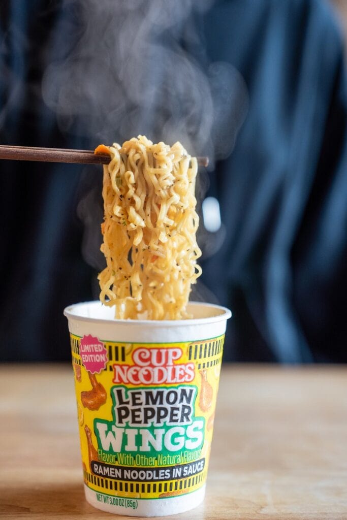 A close-up noodle pull from the Nissin Cup Noodles Lemon Pepper Wings Ramen Noodles cup with wooden chopsticks lifting a tangle of steaming yellow wavy noodles coated in pale sauce with visible black pepper flecks, the Lemon Pepper Wings cup visible below.