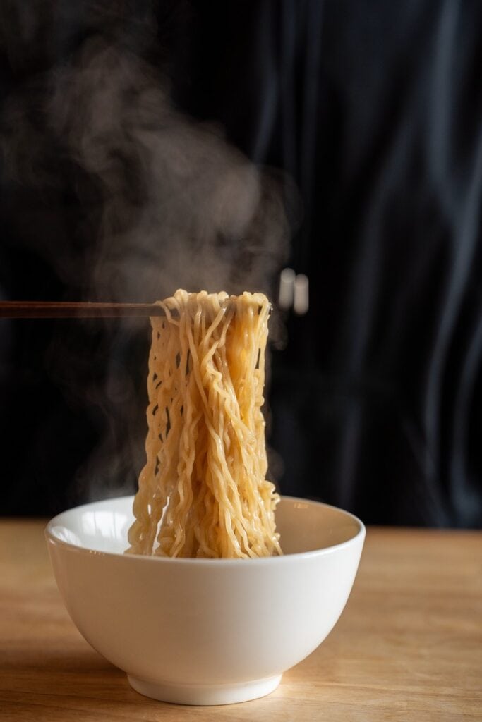 Noodle pull from Maruchan Roast Beef Ramen, showing golden wavy noodles lifted on chopsticks with steam rising above a white bowl