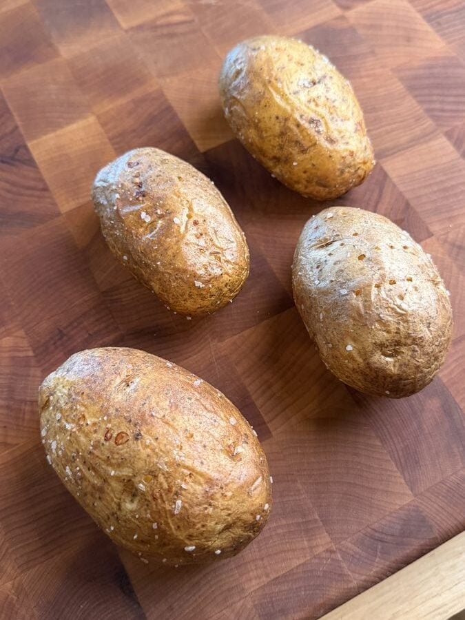 Four whole baked russet potatoes on a butcher block, skins blistered and crusted with salt, before being halved and scooped.