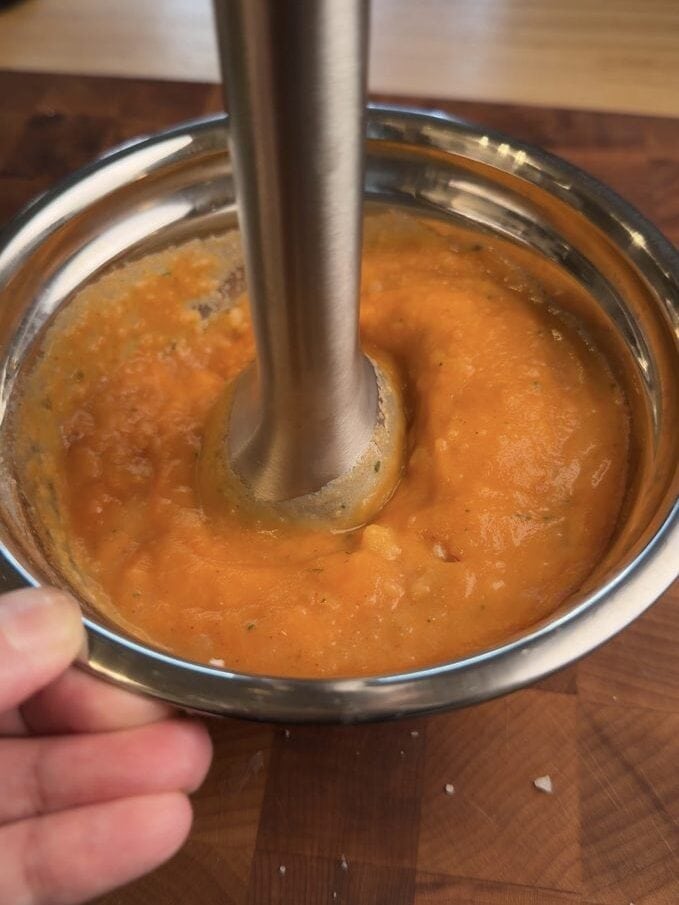 An immersion blender in a metal bowl of thick orange sauce made from the potato and ramen seasoning.