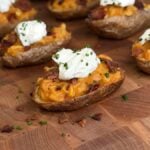 Loaded ramen baked potato half on a butcher block, filled with orange sauced instant noodles and crumbled bacon, topped with a spoonful of sour cream and sliced chives, with more halves behind it.