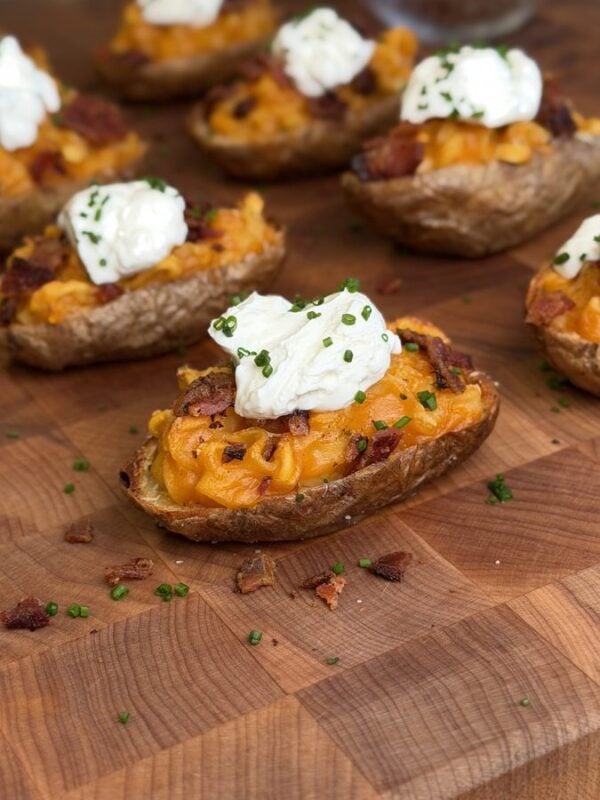 Loaded ramen baked potato half on a butcher block, filled with orange sauced instant noodles and crumbled bacon, topped with a spoonful of sour cream and sliced chives, with more halves behind it.
