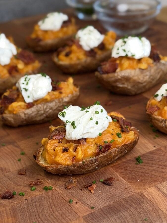 Loaded ramen baked potato half on a butcher block, filled with orange sauced instant noodles and crumbled bacon, topped with a spoonful of sour cream and sliced chives, with more halves behind it.