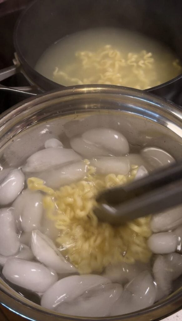 Cooked instant ramen noodles being moved with tongs into a bowl of ice water, with the noodle pot behind.