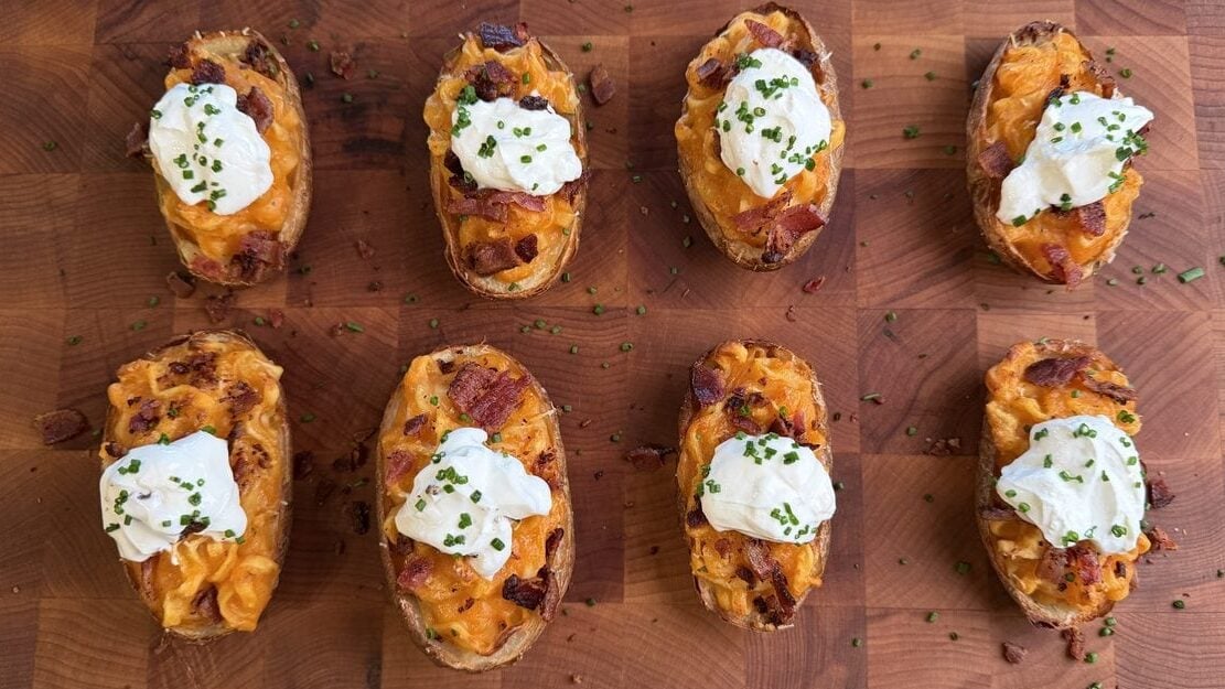 Eight loaded ramen baked potato halves arranged in two rows on a butcher block, each filled with orange noodles and bacon and topped with sour cream and chives.
