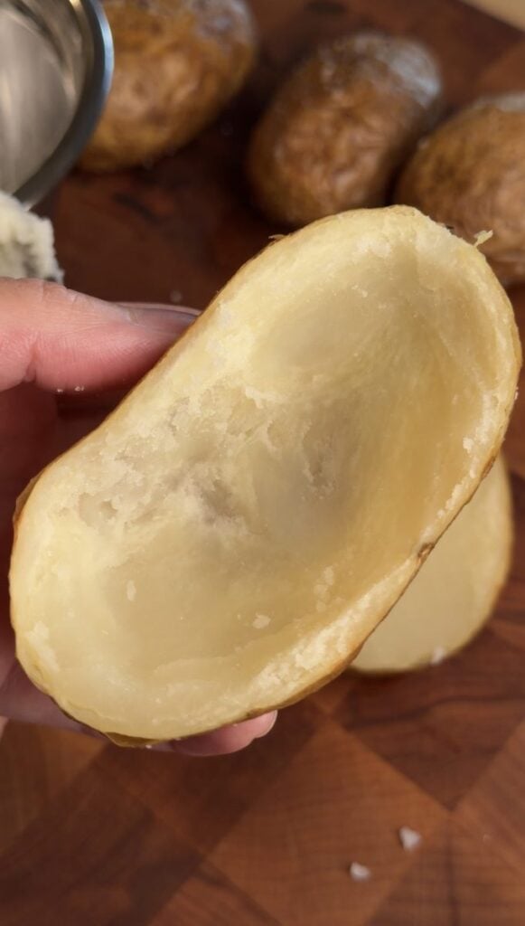 A hand holding a hollowed-out baked potato half, showing the thin wall of potato left inside the skin.