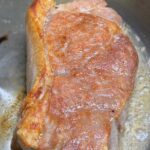A thick pork chop searing in a stainless steel pan