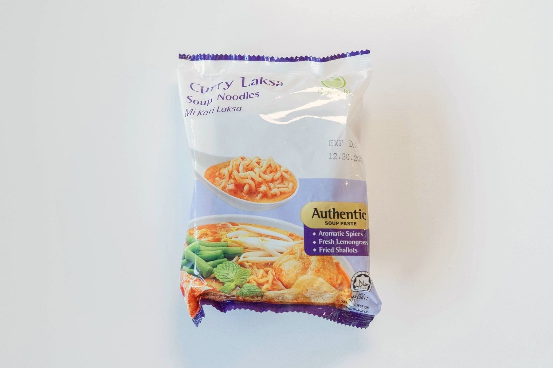 The white and purple package of KampongKu Curry Laksa Soup Noodles on a white surface, showing Curry Laksa Soup Noodles in purple script at the top with Mi Kari Laksa underneath, the small green KampongKu palm-tree logo, a photo of finished laksa noodles on a spoon and a fuller laksa bowl with snow peas and fried tofu, and a yellow Authentic Soup Paste badge calling out aromatic spices, fresh lemongrass, and fried shallots.