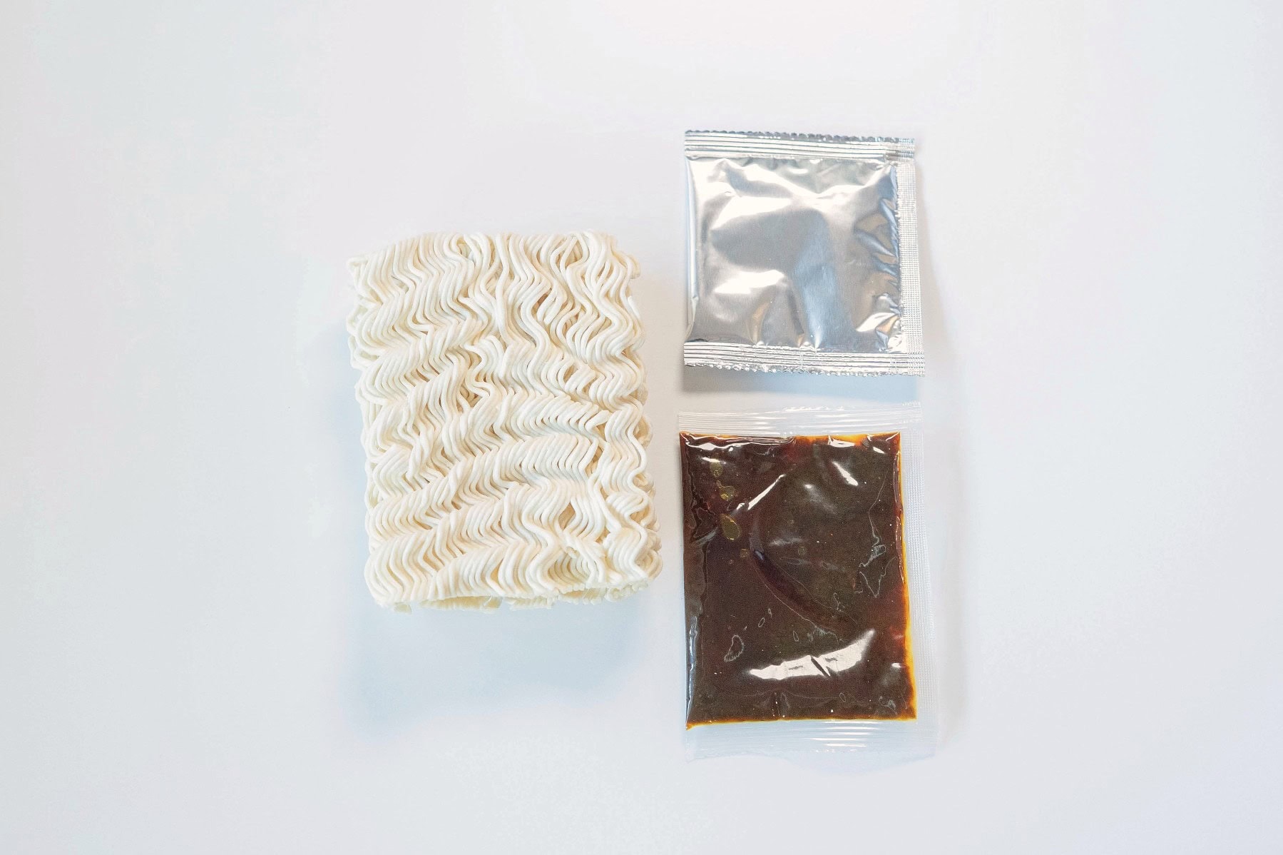 The contents of the KampongKu Curry Laksa Soup Noodles pack laid out on a white surface, showing a brick of fried wavy wheat noodles on the left, a small silver foil seasoning powder packet on the top right, and a clear pouch of thick dark brown laksa soup paste on the bottom right.