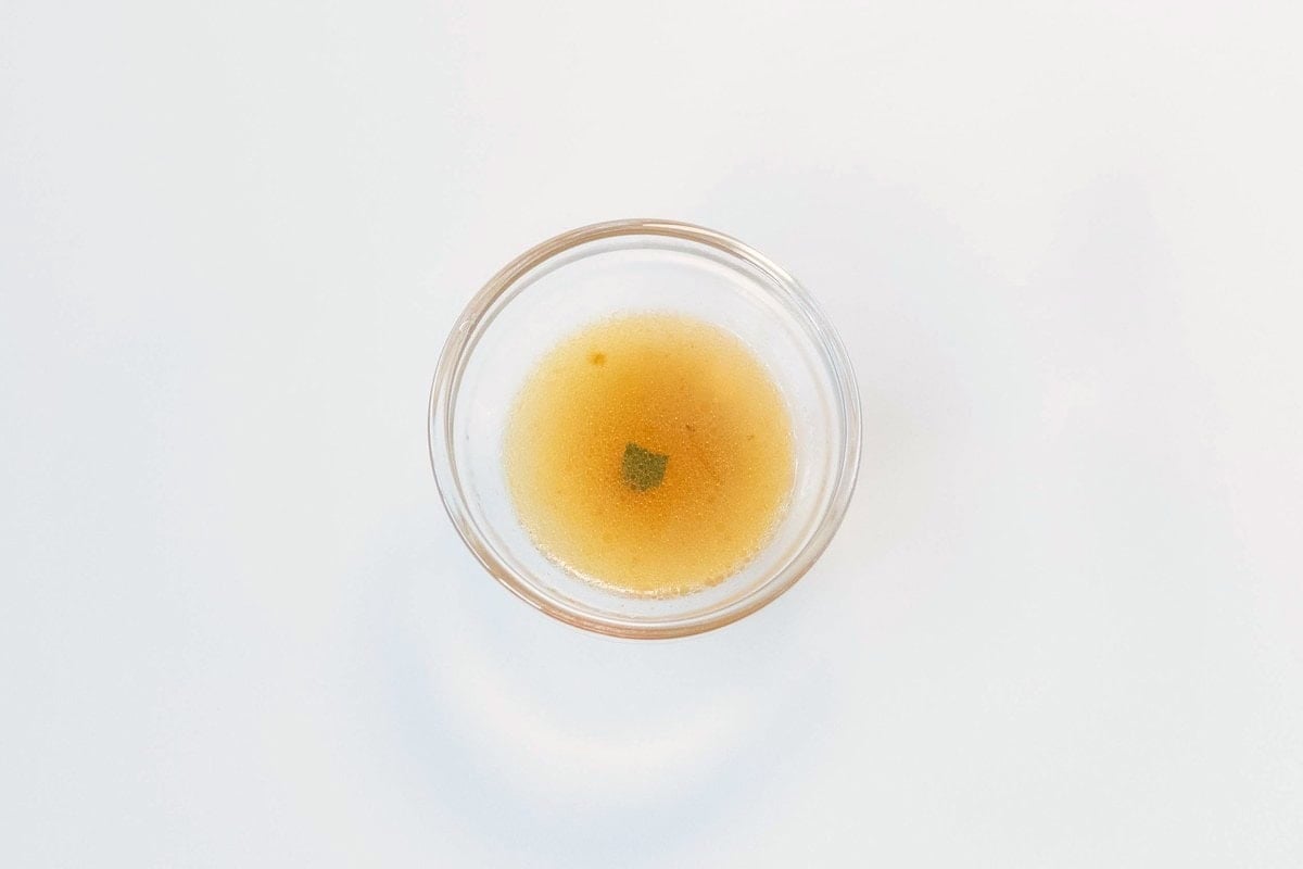 A small clear glass ramekin of the Kang Shi Fu Pork Instant Noodles with Shallots broth on a white surface, showing a clean amber-orange liquid with a slight cloudy hint and a small green herb fleck.